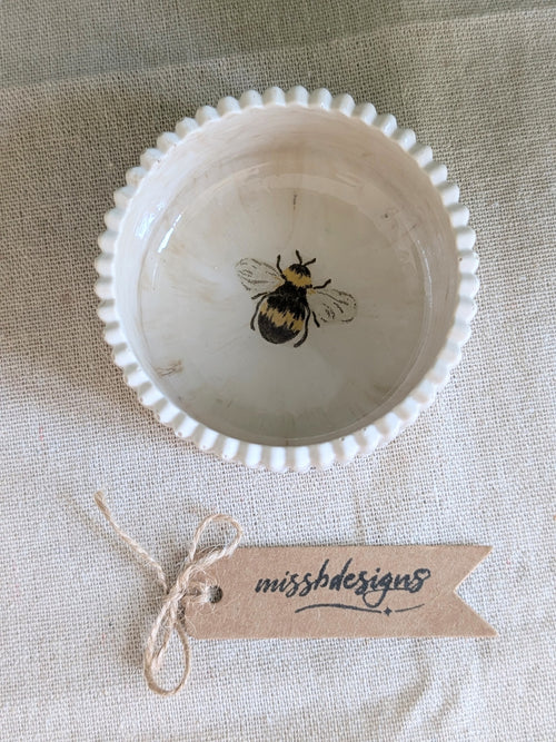 A small neutral beige ceramic ring trinket dish with a scalloped rim and a bumble bee image in the center
