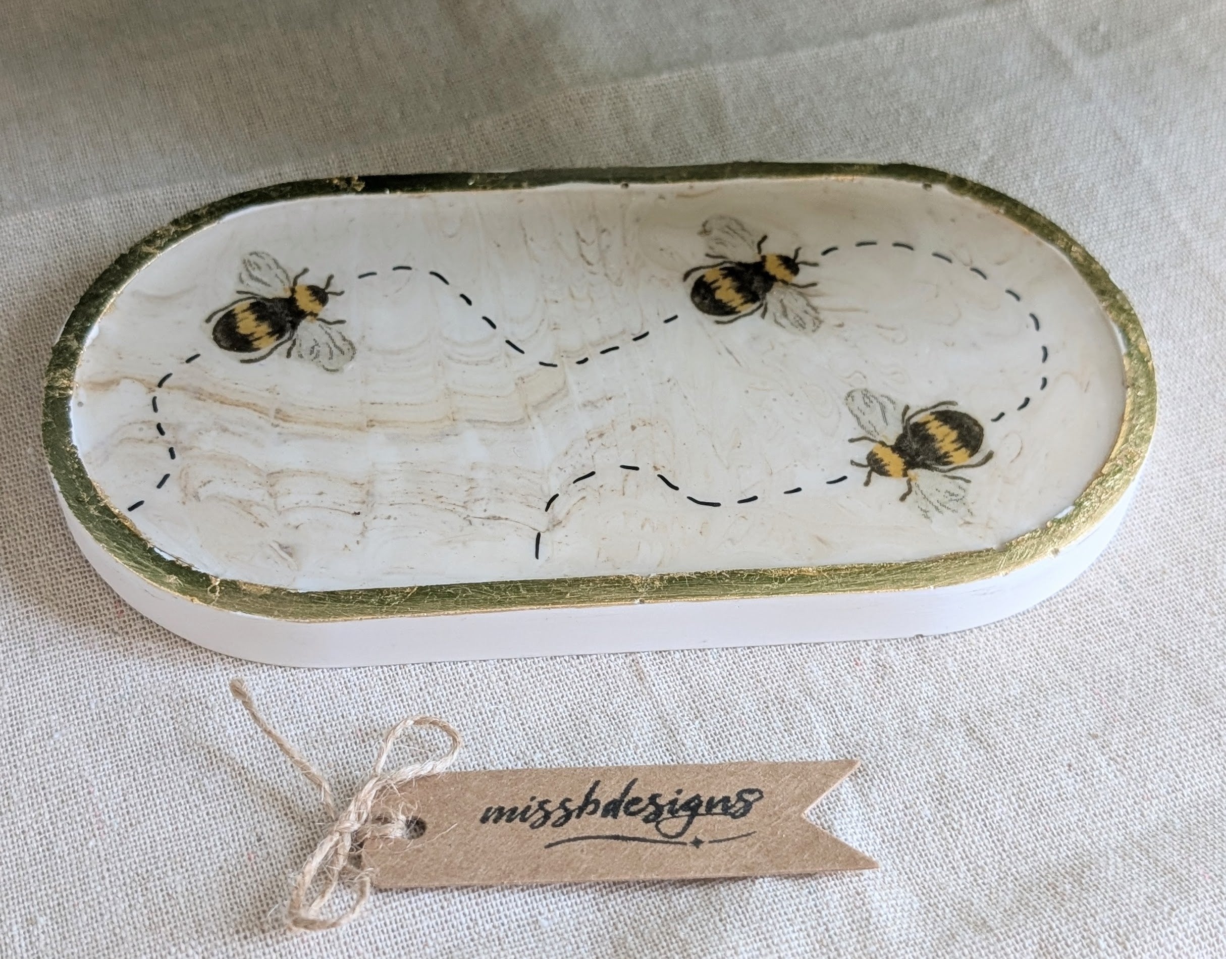 An oval ceramic dish with a distressed white glaze, featuring painted bumblebees and dashed flight paths, with a gold-toned rim. A small paper tag reading 'missbdesignts' is tied below the dish on a textured beige fabric background.