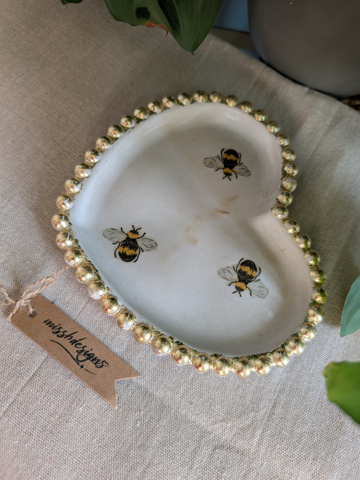 A heart-shaped white ceramic dish with a gold leaf rim and  bumblebee motifs, displayed on a beige linen surface with a brand tag attached.